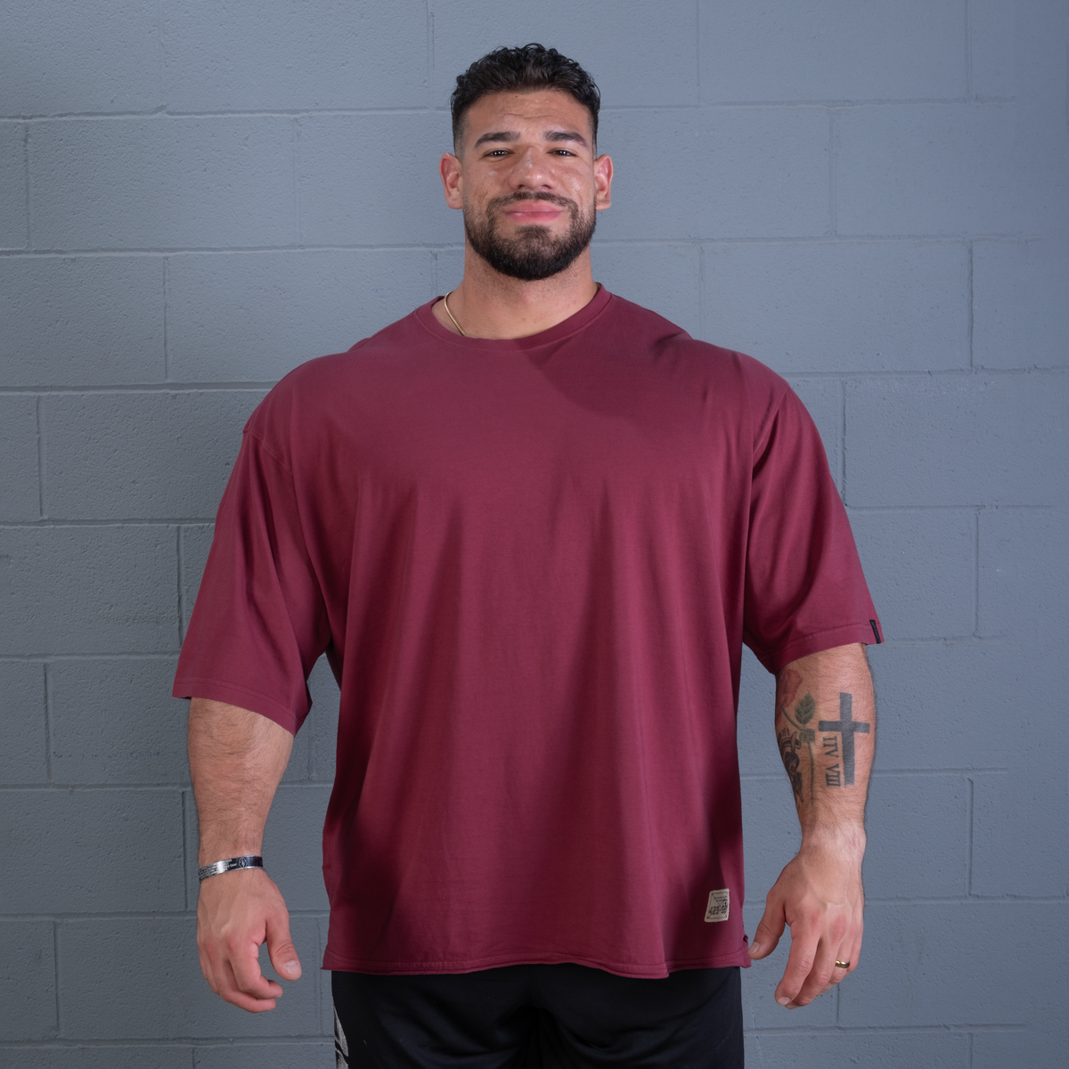 A man wearing an oversized short-sleeve crewneck t-shirt in maroon, standing against a gray painted brick wall.