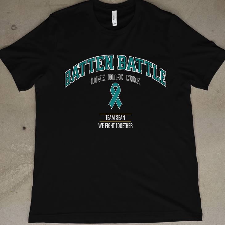 Black t-shirt with 'Batten Battle' text and teal ribbon on a concrete floor.